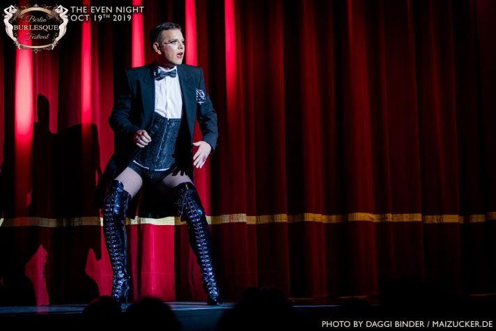 Dave van der Wal staat op het podium tijdens het Berlin Burlesque Festival 2019. Hij draagt een zwart jasje, een corset, netkousen en hoge laarzen met veters. Op de achtergrond is een rood theatergordijn te zien. Zijn expressieve houding en opvallende kleding benadrukken zijn rol als performer.