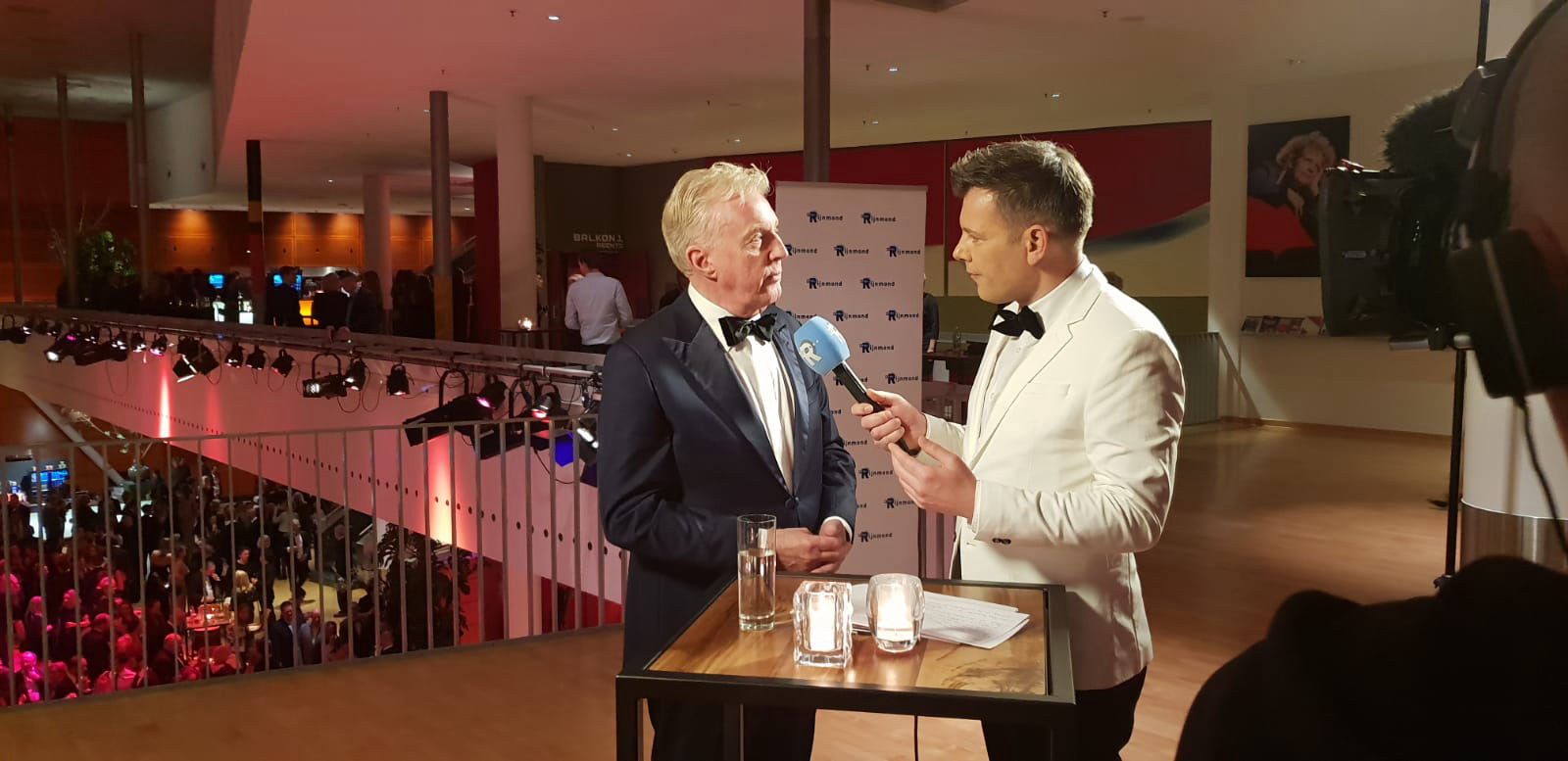 MobEen presentator in een wit pak interviewt André van Duin, die een zwart smoking draagt, tijdens de Amateur Musical Awards. Ze staan op een balkon met een drukke zaal vol mensen op de achtergrond.irise Website Builder