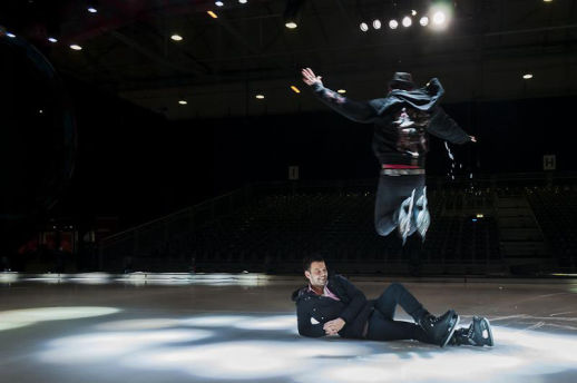 Dave van der Wal ligt op het ijs terwijl hij als reporter te gast is bij Holiday On Ice. Een schaatser springt hoog over hem heen tijdens een uitvoering. De scène is verlicht met podiumlichten, en op de achtergrond zijn lege tribunes zichtbaar. Dave lacht terwijl hij het spectaculaire moment vastlegt.