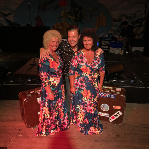 Dave van der Wal poseert samen met de accordeonisten, de zusjes van Wanrooy. Ze staan op een podium, waarbij Dave in het midden staat en een arm om elke zus heeft geslagen. De zusjes dragen identieke kleurrijke jurken met een bloemenpatroon. Achter hen is een decoratief schilderij en muziekapparatuur te zien. De sfeer is gezellig en ontspannen.