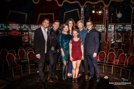 Dave van der Wal, tweede van links, staat in de Spiegeltent in Rotterdam tijdens het tweede jaar van de eigen productie Wintervarieté. Hij is omringd door cast en crew. Helemaal rechts staat David Severins, mede-producent. De groep staat in een sfeervolle omgeving met gekleurde ramen en lege stoelen op de achtergrond.