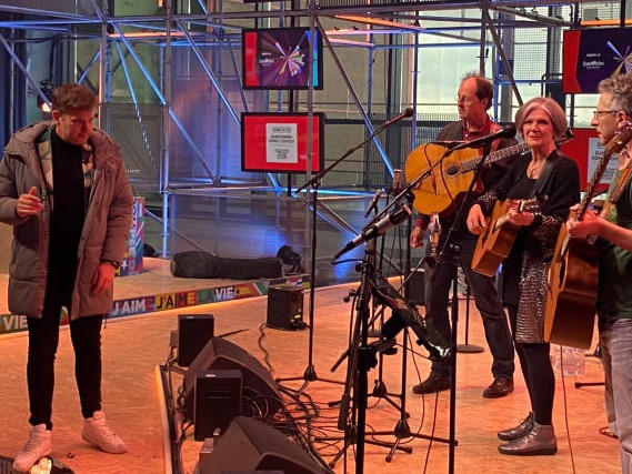 Dave als redacteur - producent tijdens de dagelijkse talkshow van het eurovisie songfestival. Hij draagt een lange grijze winterjas, staat in de studio terwijl zangeres Lenny Kuhr aan het repeteren is voor haar nummer de troubadour