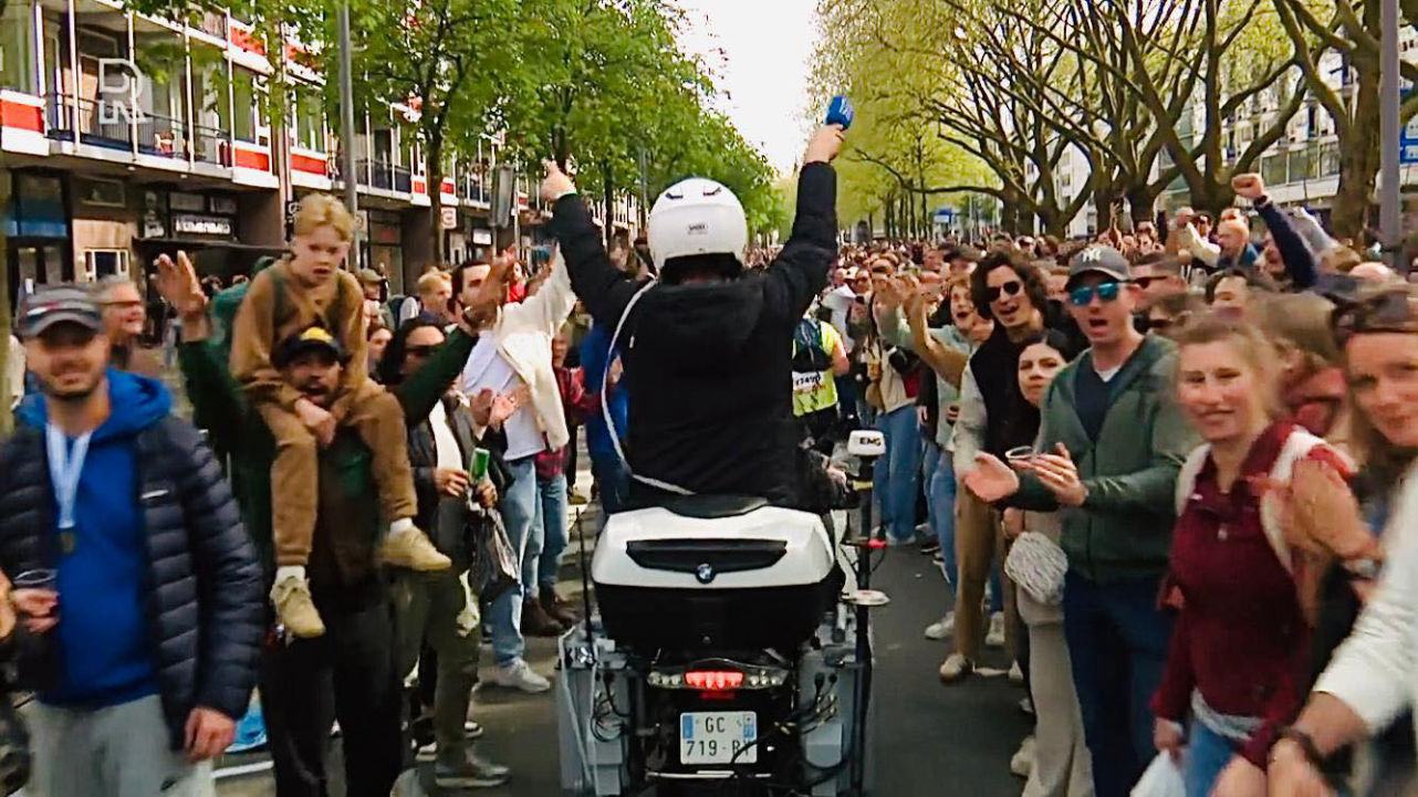 Dave van der Wal zit als verslaggever achterop de motor tijdens de Rotterdamse Marathon. Hij draagt een witte helm en steekt zijn armen in de lucht terwijl hij door een enthousiaste menigte rijdt. De straat is gevuld met toeschouwers die juichen en applaudisseren tijdens het evenement. Beeld: RTV Rijnmond