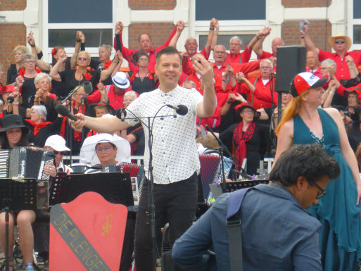 Dave van der Wal, als vaste presentator tijdens het O Festival in Rotterdam, leidt de productie "Van Smartlap tot Opera". Hij staat voor het orkest, gekleed in een wit overhemd met stippen, terwijl hij enthousiast het publiek toespreekt. Achter hem zingt een groot koor in rode en zwarte kleding. De sfeer is levendig en feestelijk.