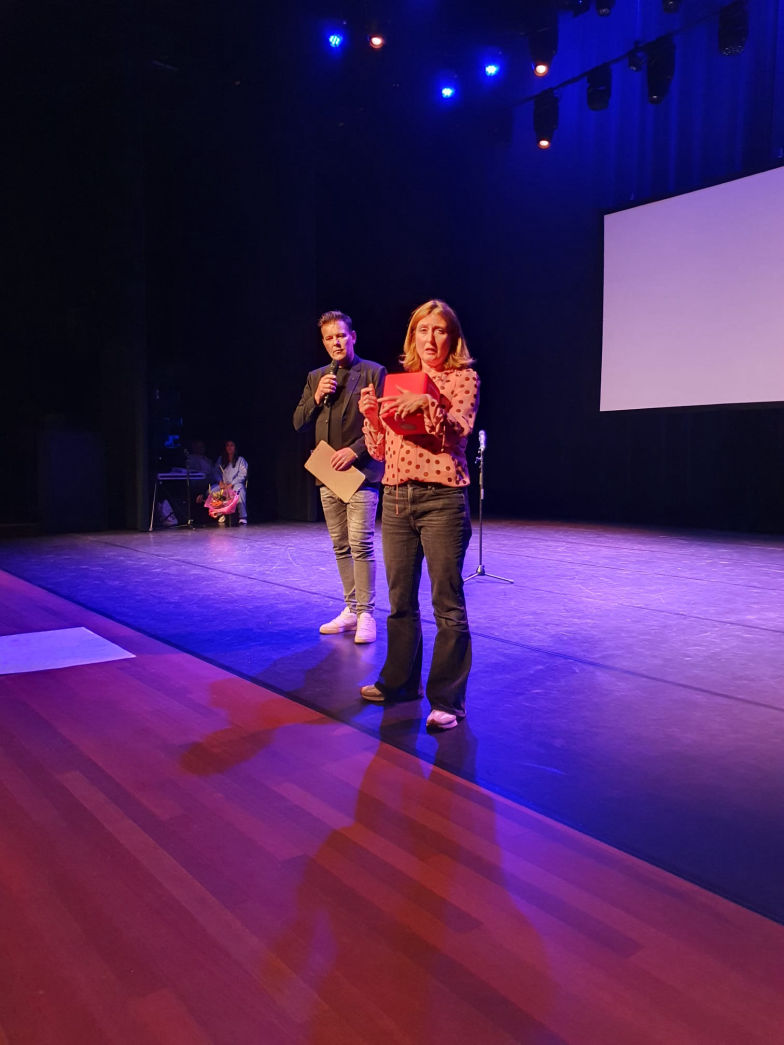 Dave van der Wal staat als dagvoorzitter op het podium tijdens een evenement van Woonstad Rotterdam. Hij draagt een donkere outfit en spreekt in een microfoon, terwijl een vrouw naast hem iets presenteert met een rood mapje in haar hand. De scène is verlicht met blauwe lichten en op de achtergrond is een leeg projectiescherm te zien.